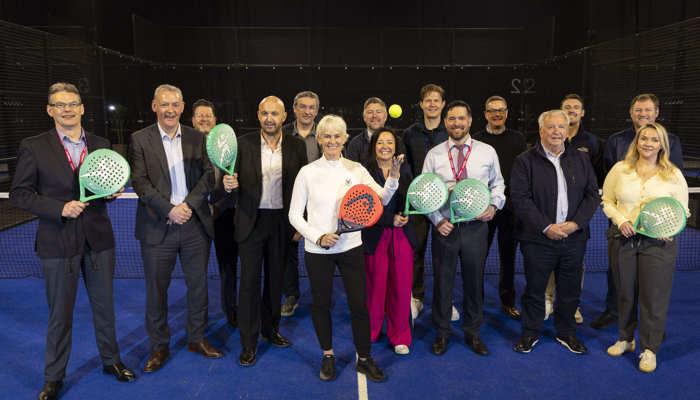 A large group of people stand on a padel court, with some holding padel racquets and one throwing a padel ball in the air
