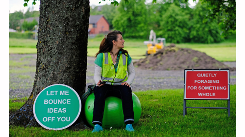 The artist sits on a green yoga ball with a high vis vest. To there left a green sign reads "let me bounce ideas off you".