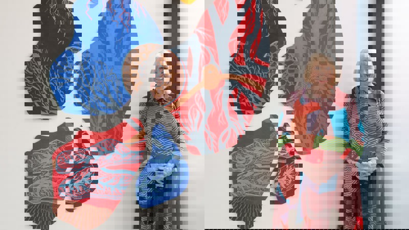 A woman stands beside a wall installation made of several irregular, organic‑shaped panels covered in intricate, vein‑ and branch‑like patterns. Each panel is painted in vivid colors—reds, blues, yellows, and greys—with fine contrasting line work. Her outfit echoes the artwork’s palette and patterns, creating a visual harmony between her clothing and the installation. The setting appears to be a bright, modern gallery space with large windows letting in natural light.