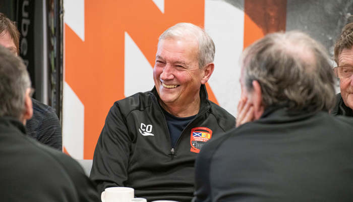 An elderly person with a big smile sits around a table with others. They have coffee cups on the table.