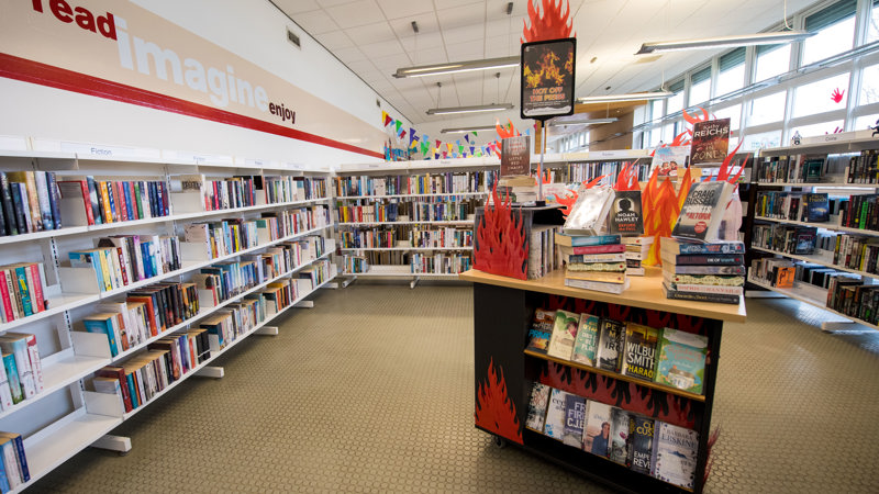 A large selection of bookshelves. There is wall writing on the left hand wall that reads 'Read, Imagine, Enjoy'