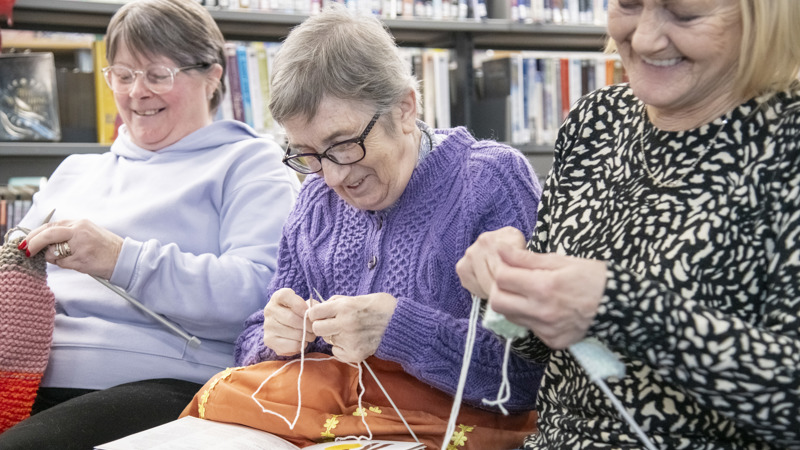 Three people smiling while they are sitting down and knitting with needles and yarn in a library.