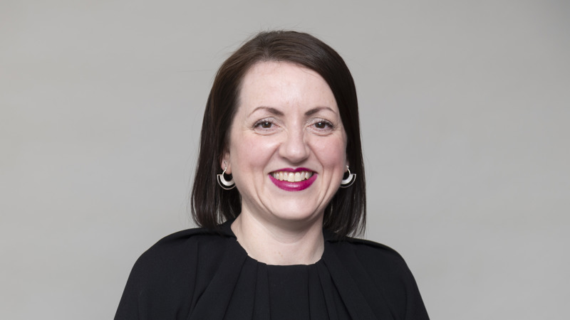 Headshot of a person with dark hair, wearing a black top, smiling, against a neutral studio background.