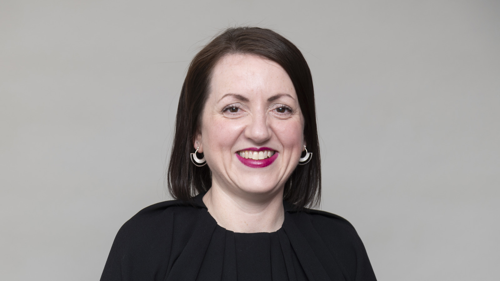 Headshot of a person with dark hair, wearing a black top, smiling, against a neutral studio background.