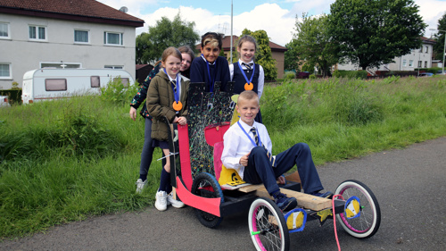 Children show off their hand made racing cart. The cart is a bright red and has three wheels.