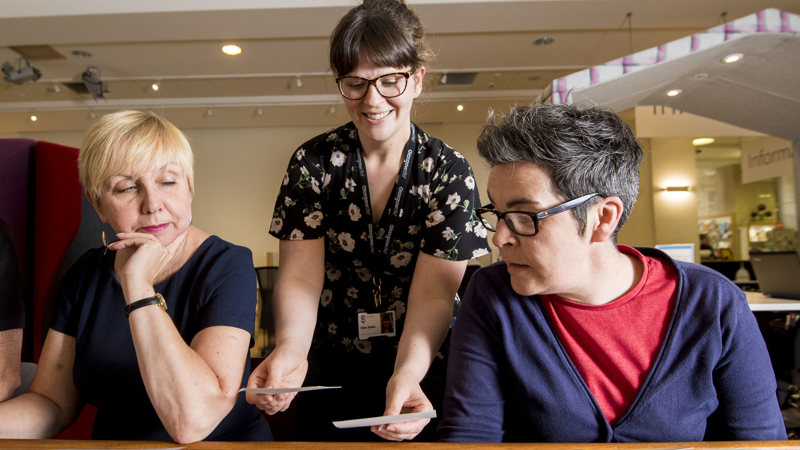Three people, two sitting at a desk and one standing, all three looking at information on the table. 
