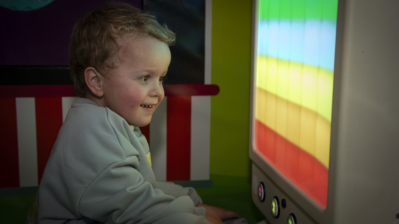 A young child with blonde hair smiling as they look at a multi-coloured sensory wall.