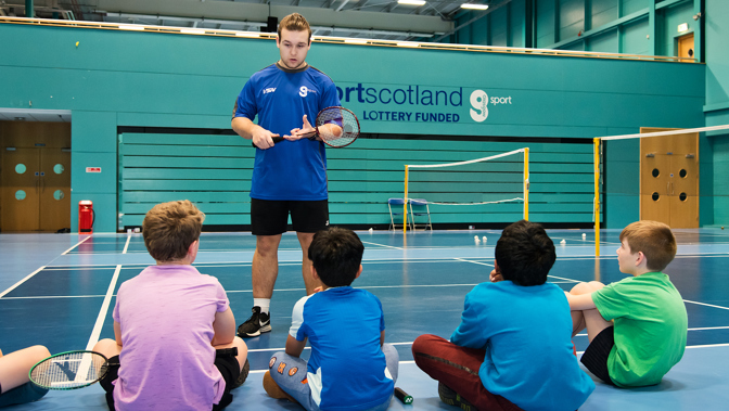 In the middle of a badminton court, a coach is standing up educating a group of young children who are sitting down on the rules of the game.