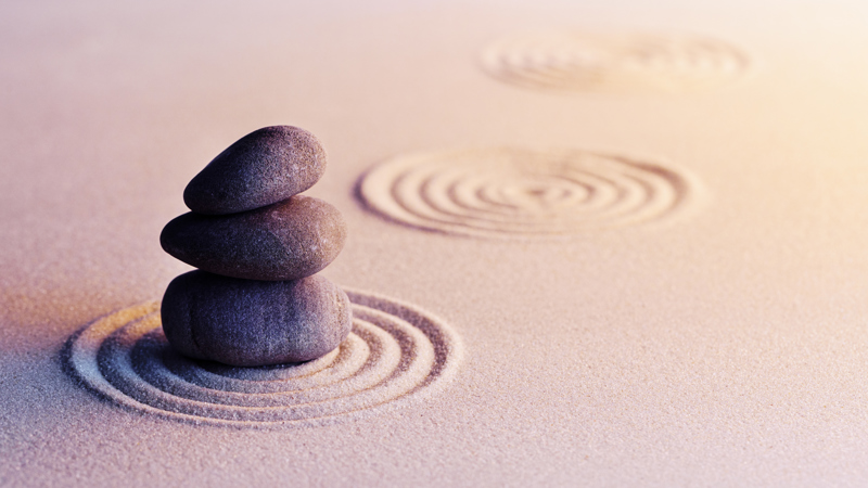 Three smooth stones stacked on raked sand, with circular ripple patterns around them in a calm, minimalist scene.