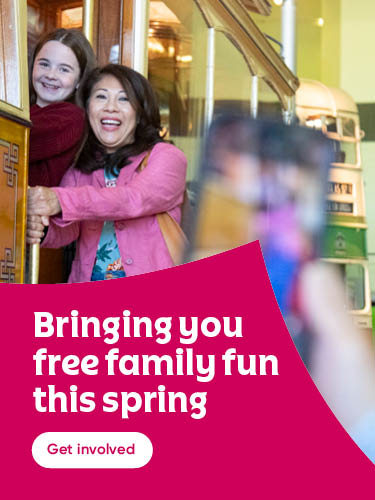 Two smiling people stand at the entrance of a vintage yellow tram inside a museum, while someone in the foreground takes their photo with a smartphone. A bright pink banner on the left reads ‘Bringing you free family fun this spring’ with a ‘Get involved’ button below.