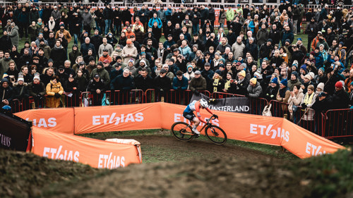A cyclist races round a muddy track cheered on by spectators