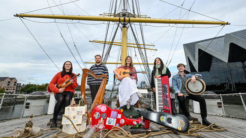Musicians sit holding a mix of instruments in front of instrument cases, rope, glass bottles and crates on board a ship