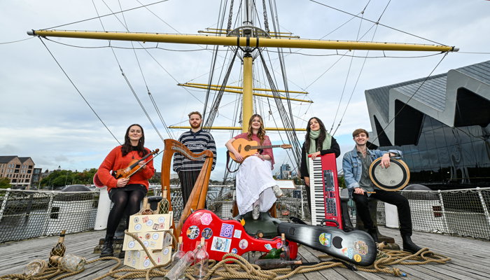 Musicians sit holding a mix of instruments in front of instrument cases, rope, glass bottles and crates on board a ship
