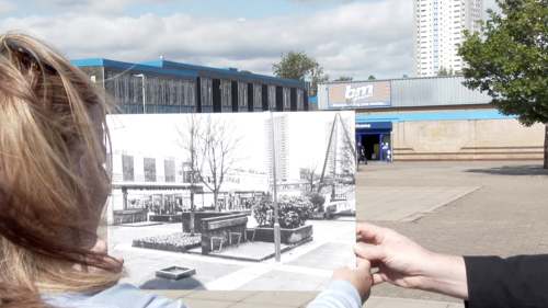 Two people look at an old photograph of the site they are looking at. The site in the past has buildings which are now demolished.