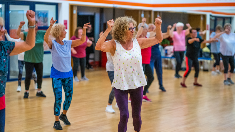 A group of women during a fitness class in an exercise hall, they all have their arms raised.