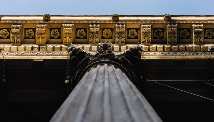 An upward view of a classical stone column with fluted sides and an ornate Corinthian capital decorated with carved acanthus leaves and scrolls. Above it, the entablature features intricate floral patterns, dentils, and layered moulding, all captured from a low angle that emphasizes the structure’s height and elaborate craftsmanship.