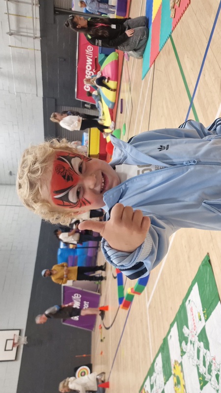 Child taking part in a fun holiday activity session, giving a thumbs up in a sports hall filled with games and play activities.