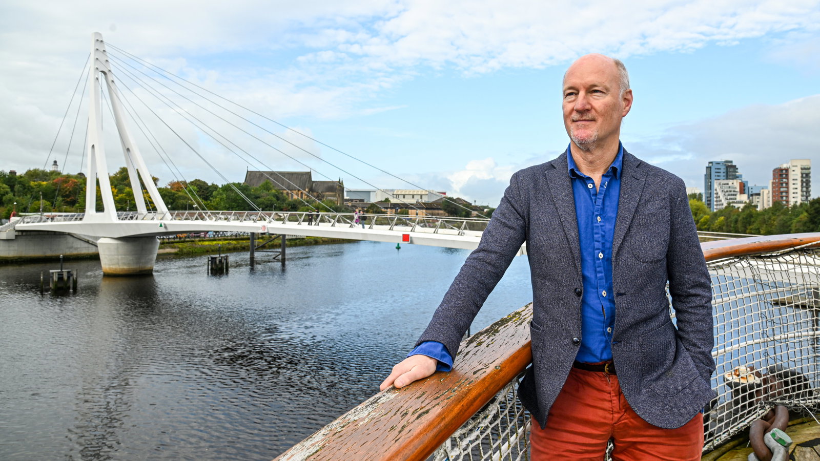A man stands on board a ship and looks into the distance with a white bridge crossing a river in the background