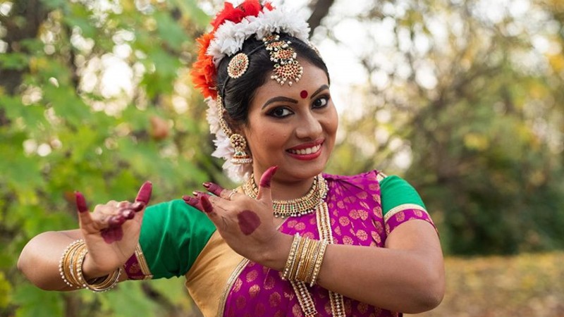 A woman in red, white, green and purple South Asian clothing is positioning her hands to her side as part of a dance routine