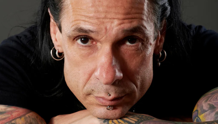A man with long dark hair and earrings is leaning forward and resting his chin on his hands