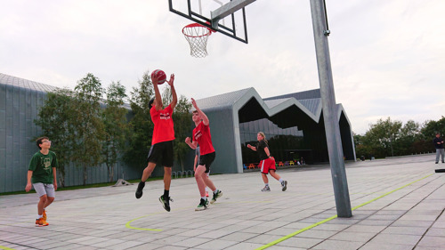 Close-up action shot of two boys jumping to shoot a basketball near the hoop, with a distinctive modern building in the background.