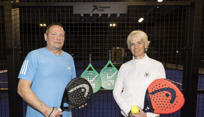 A man and a woman stand beside one another while holding padel racquets in front of jacket hooks and a sign for 'Glasgow Club'