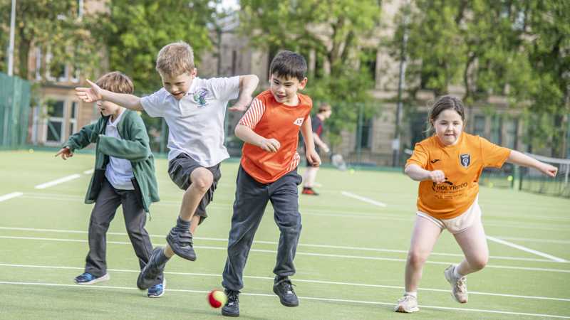 Four children are mid motion, running and leaping, after a tennis ball on a green tennis court. 