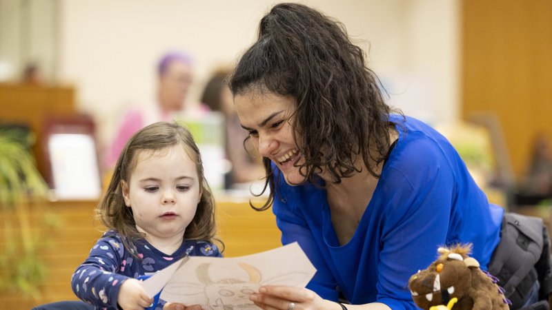 And adult helping a child cut out a paper outline of the Gruffalo in a library, there are colourful pens as well as Gruffalo soft toy on the table next to them.
