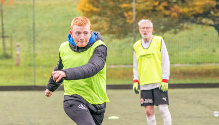 A younger person strikes a football, while an older person behind watches on.