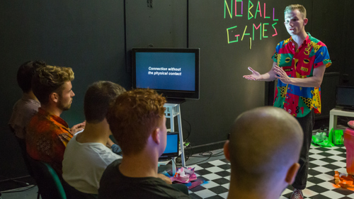 A person, is performing a play they are wearing a tropical shirt and pointing to a TV. In the background is neon tape that reads "No Ball Games".