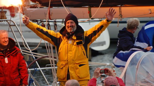 A person wearing a yellow waterproof jacket is smiling and  holding a celebratory flare on the deck on a yacht 