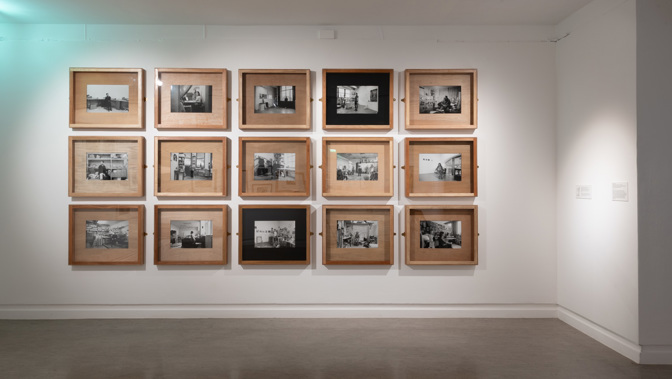 A gallery wall displaying fifteen black‑and‑white photographs arranged in three rows of five, each in a wooden frame. The images depict a mix of indoor and outdoor scenes, including people in everyday environments, architectural interiors, and street views, creating a cohesive documentary-style collection