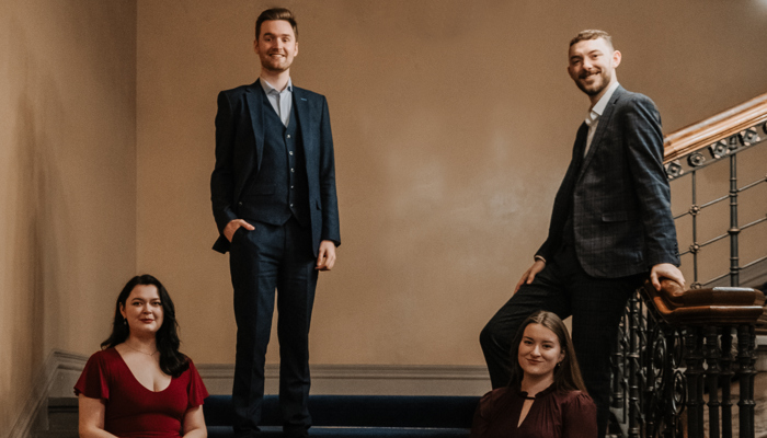 Four people smiling directly at the camera, two are sat on some stairs and the other two are stood.
