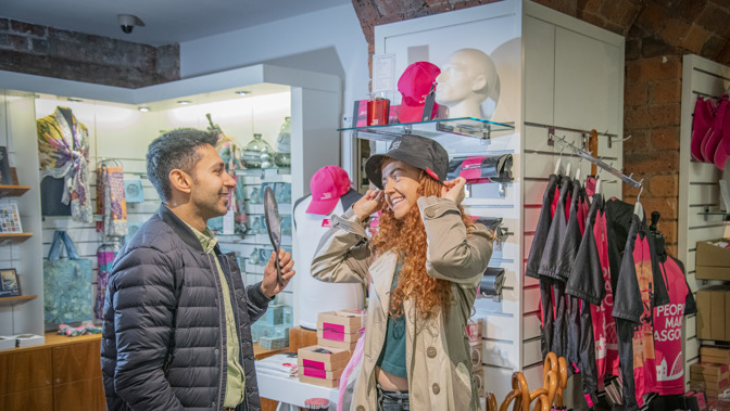 Two people stand inside a Glasgow Life museums gift shop. A woman with long curly hair smiles while trying on a black cap, adjusting it with both hands. A man beside her, wearing a dark jacket, holds up a handheld mirror so she can see herself. Shelves around them display colourful hats, scarves, umbrellas, and other merchandise, with bright pink items prominently featured.