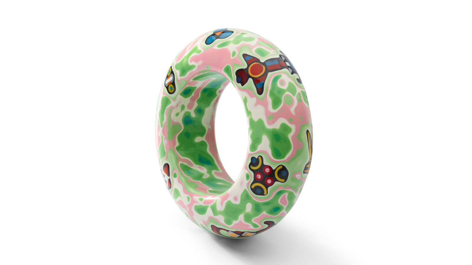 Decorative torus-shaped sculpture with a smooth, glossy surface featuring swirling pink, green, and white marbled patterns. The object is embellished with colorful geometric motifs in red, blue, yellow, black, and white, and is displayed against a plain light grey background with a soft shadow beneath it.
