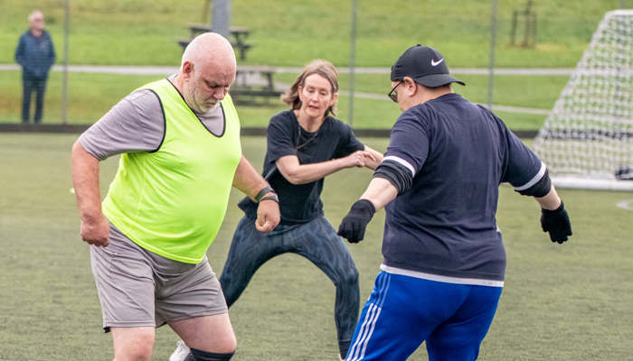 Three footballers go in for the ball, which is between them on an outdoor pitch.