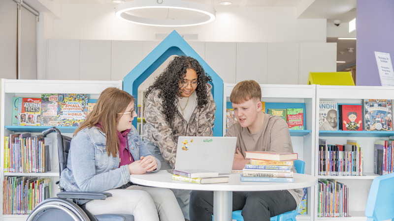 A group of three people sit together at a round table in a brightly lit library. One person is using a wheelchair, another is seated on a chair, and a third person stands behind them. They are gathered around an open laptop with books stacked nearby. Shelves filled with colorful children's books surround the group.