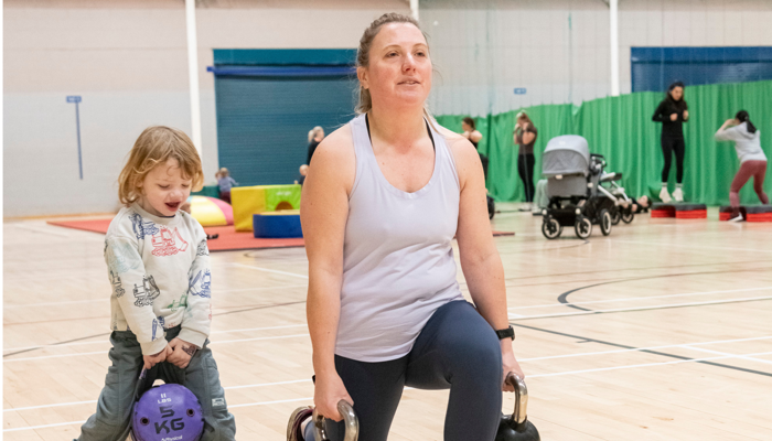 A parent does a lunge exercise holding kettlebells in a sports hall, while a young child stands nearby holding a small weight, copying the movement.