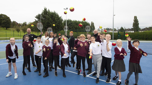 School children at Garrowhill Park tennis courts throwing tennis balls towards the camera with adults holding rackets up behind them.