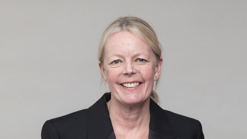 Headshot of a person with light hair tied back, wearing a dark blazer, smiling, against a neutral background.