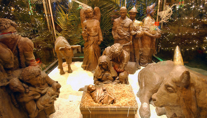 A detailed nativity scene display featuring statues of Mary, Joseph, baby Jesus in a manger, animals, and other figures, surrounded by straw and illuminated by warm lights.