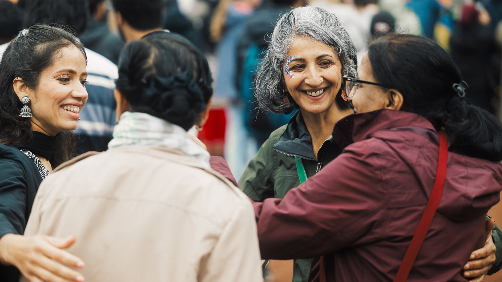 A group of four people are smiling and dancing together at an outdoor festival event. They are wearing rain jackets 