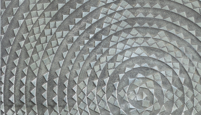 Close-up of a textured grey surface engraved with concentric spiral bands made up of small triangular and diamond-shaped motifs. The geometric pattern radiates inward toward a central spiral, creating a sense of movement and depth. The matte, stone-like surface shows subtle variations in tone and texture.