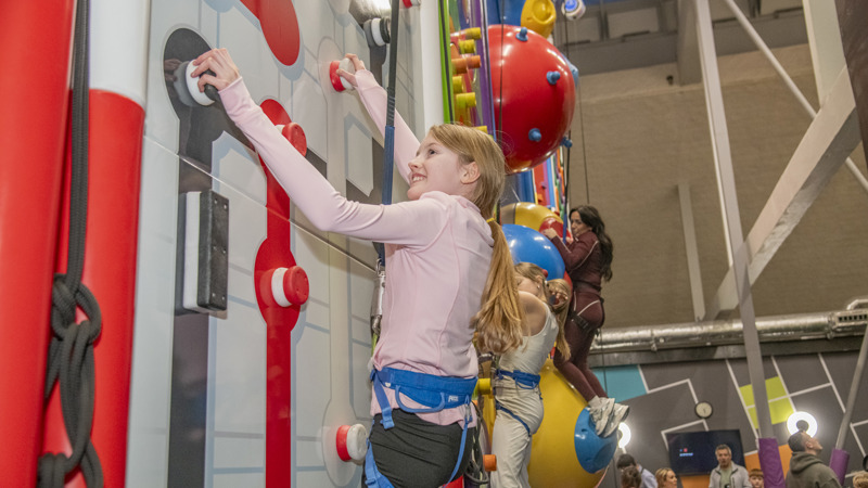 A smiling young girl with a pink zipper and blue harness on climbing up a climbing wall.