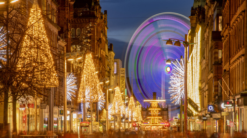Glasgow’s Buchanan Street decorated with festive Christmas lights at night. Golden tree-shaped lights and bright star decorations line both sides of the street, while a large, colourful Ferris wheel glows in the background. 