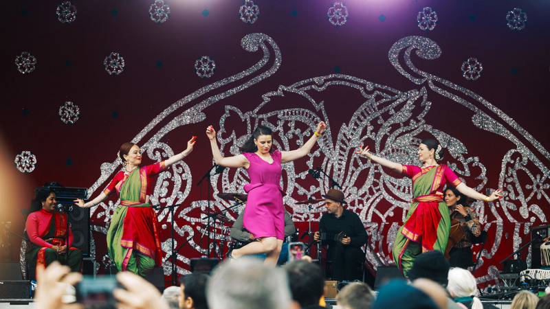 Three performers dance on a stage with musicians behind them and a large red and silver decorative backdrop, as a crowd watches.