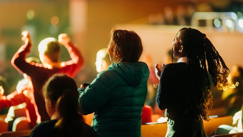 School children seen from behind, cheering and clapping during a concert in a dimly lit hall with warm stage lighting.