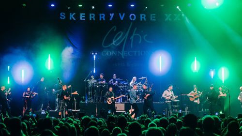 A large crowd is watching several musicians and singers perform on a colourfully lit stage