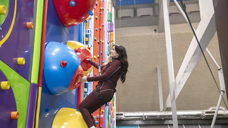 A woman with black hair in maroon gym leggings and zipper climbing a multi-coloured Clip 'n Climb wall