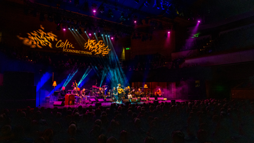 Musicians on a stage with multicoloured stage lights. The words Celtic Connections are displayed in the concert hall.